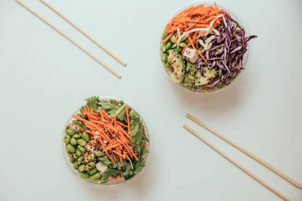 Twee poke bowls met wortels en groentes met stokjes op een witte tafel.