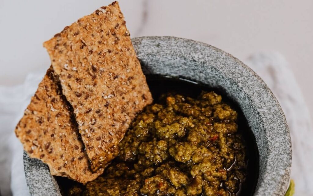 Koolhydraatarme crackers met gemixte zaden en pitten in een schaaltje met tapenade op een witte achtergrond.